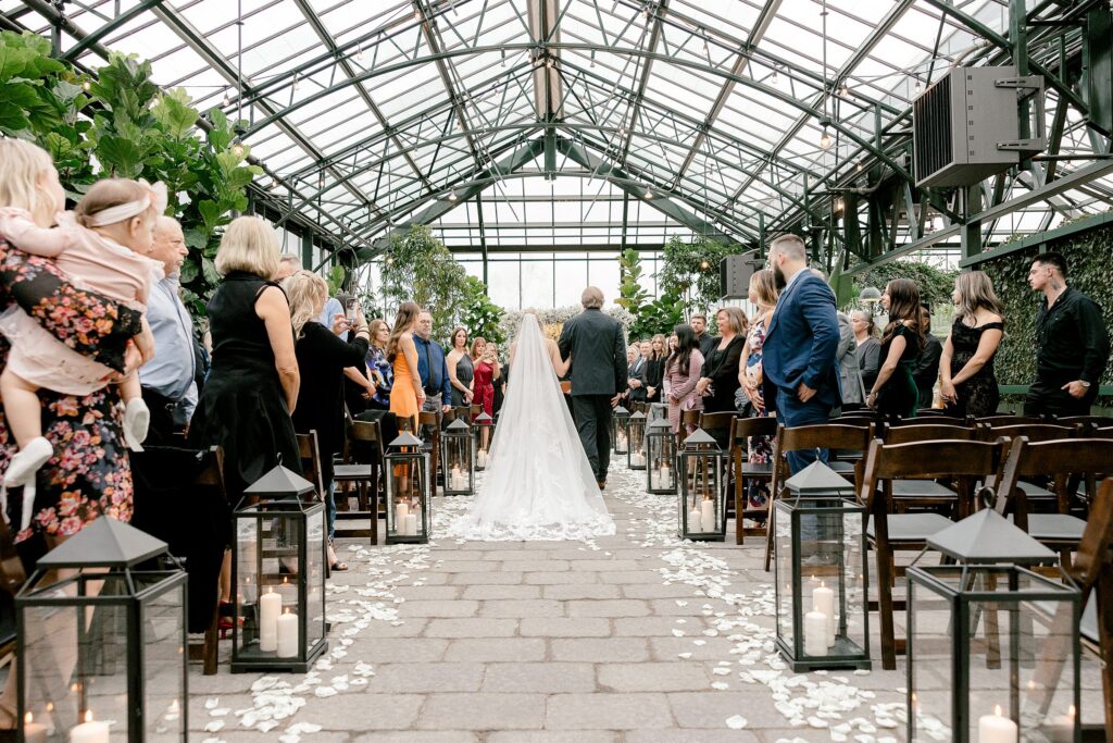 bride being walked down the aisle inside greenhouse wedding venue
