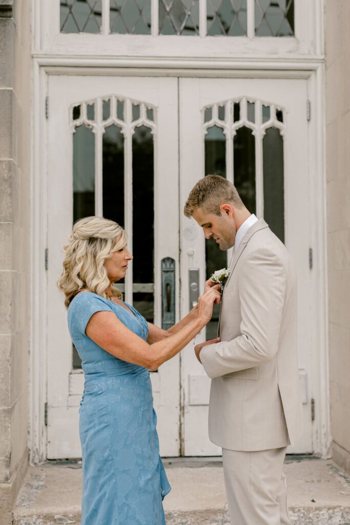 Groom's mom pinning on his boutonniere in front of the white church doors at The Broadway Avenue in Grand Rapids