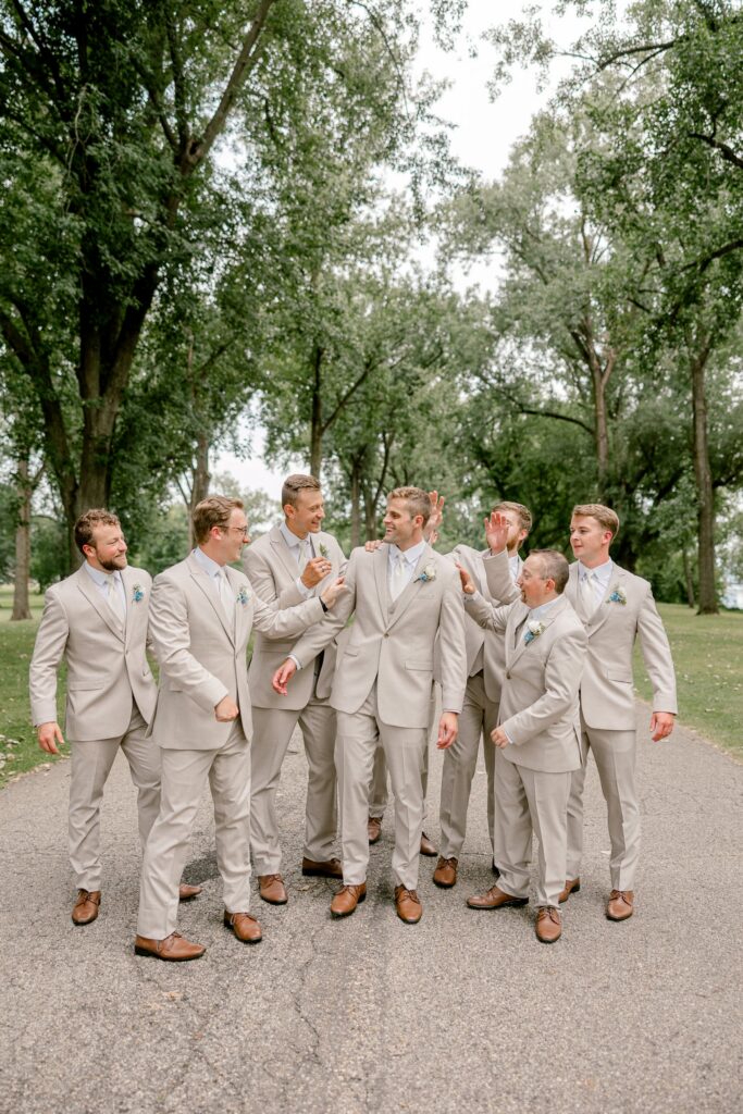 Groom laughing with his groomsmen in tan suits on a tree-lined path at Riverside Park in Grand Rapids