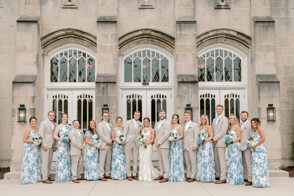 Full wedding party in blue floral dresses and tan suits in front of the arched stone entrance of The Broadway Avenue in Grand Rapids
