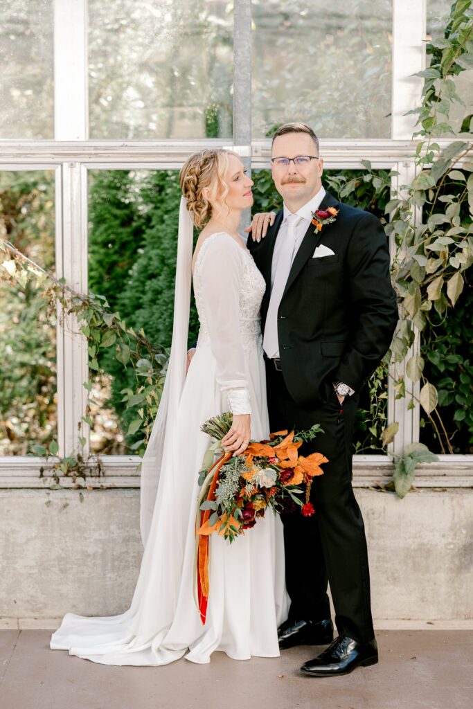 Bride and groom standing by the greenhouse windows with greenery behind them
