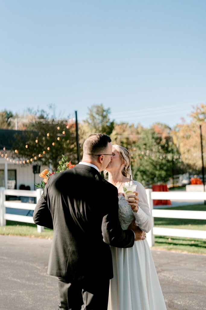 Bride and groom sharing a kiss with drinks in hand by the white fence