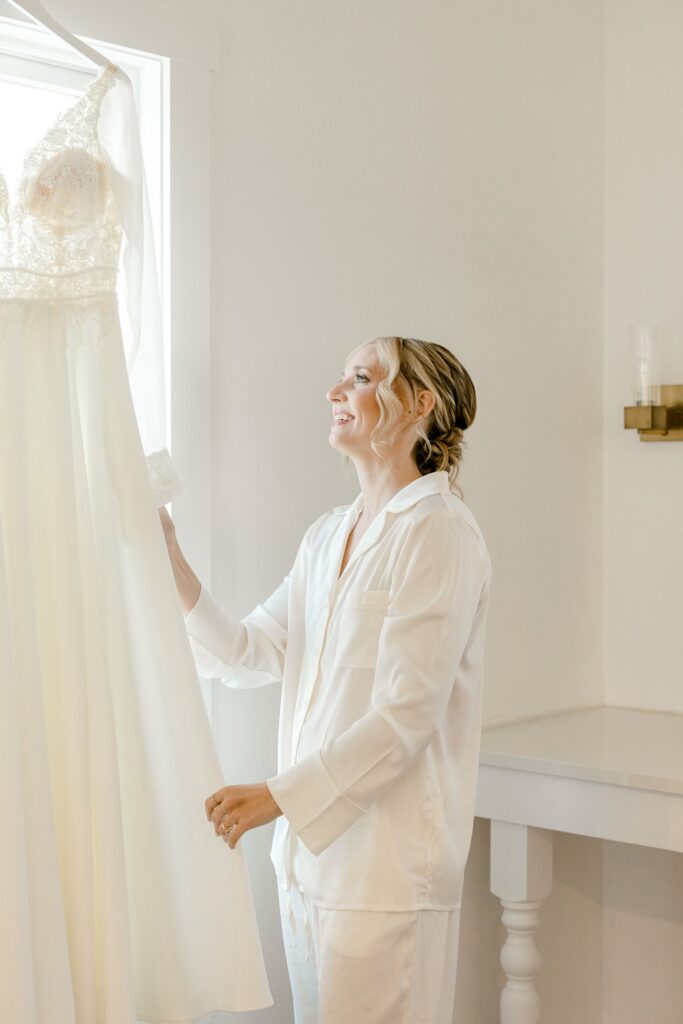 Bride in a white robe looking up at her wedding dress by the window