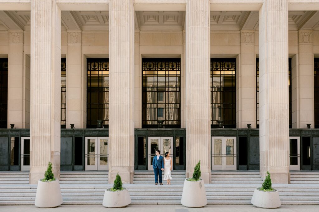 Engaged couple standing on the steps between the tall limestone columns of the Civic Auditorium in downtown Grand Rapids