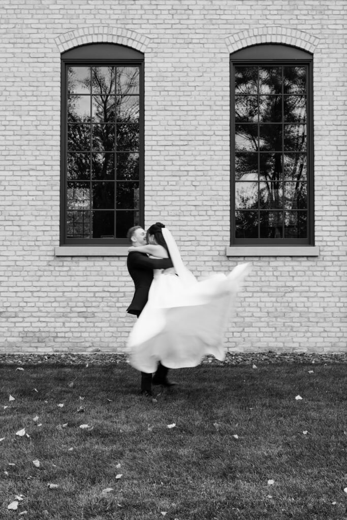 Black and white photo of the groom lifting the bride by the brick windows