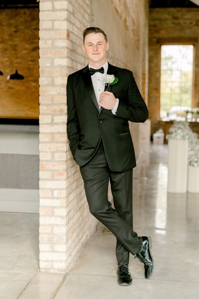 Groom in a black tuxedo leaning against a brick wall at The BRIX on Fox