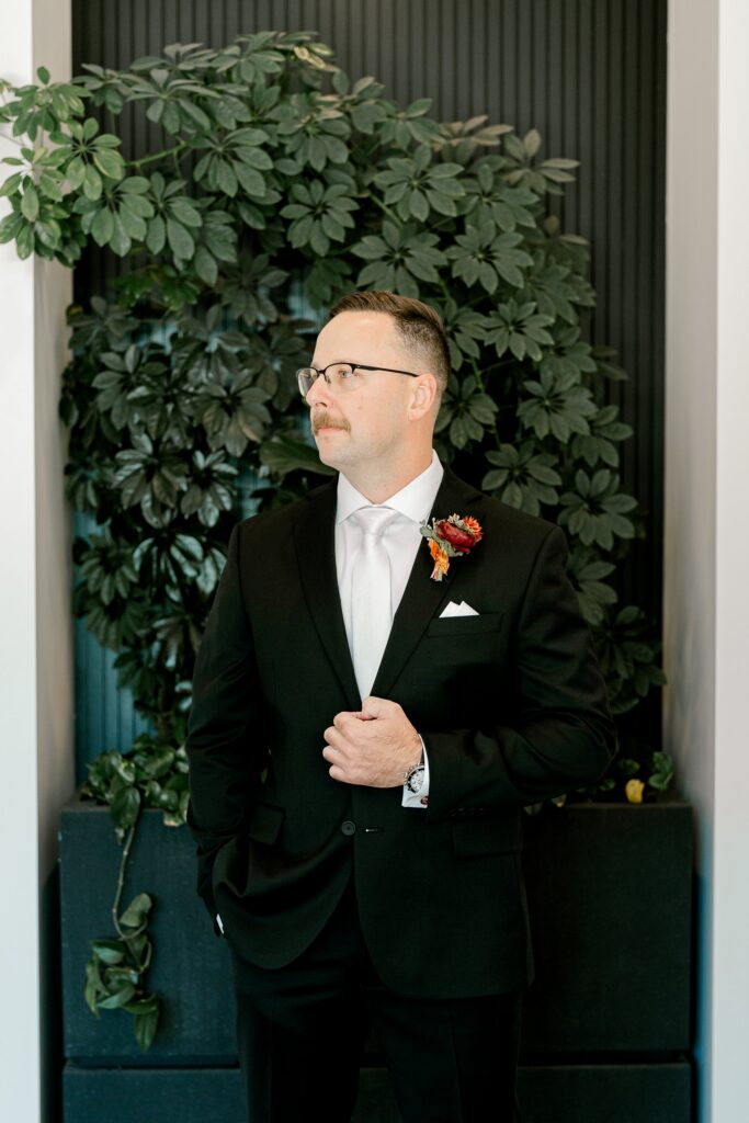 Groom in a black tux with a rust boutonniere in front of a greenery wall