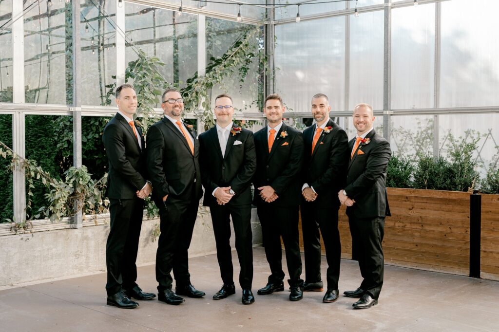 Groom and groomsmen in black tuxes with rust ties inside the greenhouse