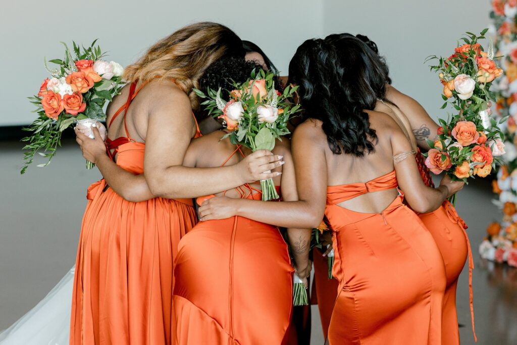 Bridesmaids in burnt orange dresses hugging the bride during their first look at Leona Road
