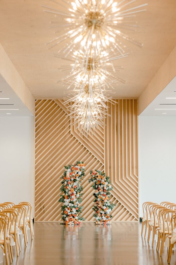 Leona Road ceremony space with a wood slat wall, orange florals and a starburst chandelier