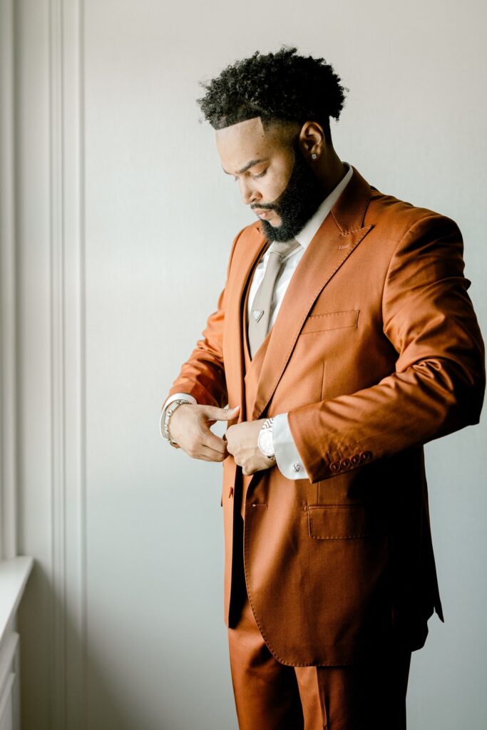Groom in a rust orange suit getting ready in window light at the Amway Grand Plaza in Grand Rapids