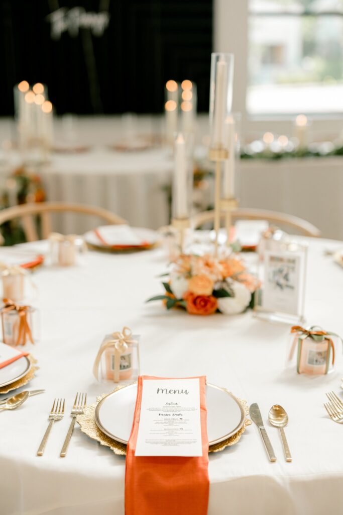 Reception place setting with an orange napkin, gold flatware and menu card at Leona Road