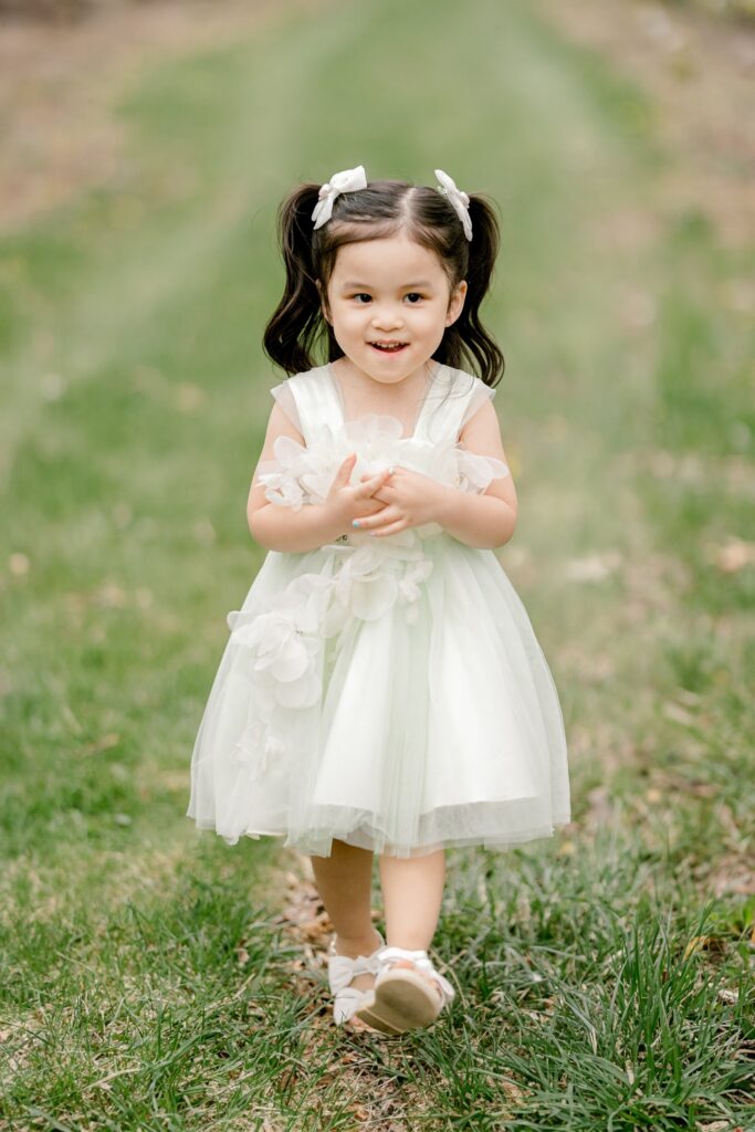 Little girl standing in the grass in her white dress during a spring orchard session in Grand Rapids