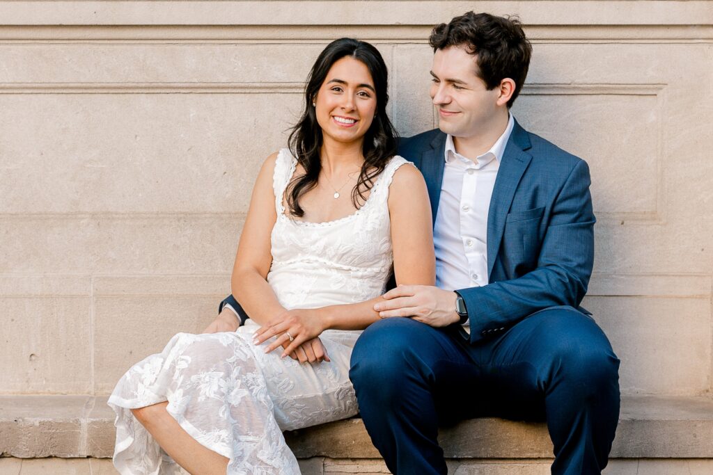 Engaged couple sitting together on a stone ledge against a historic building in downtown Grand Rapids