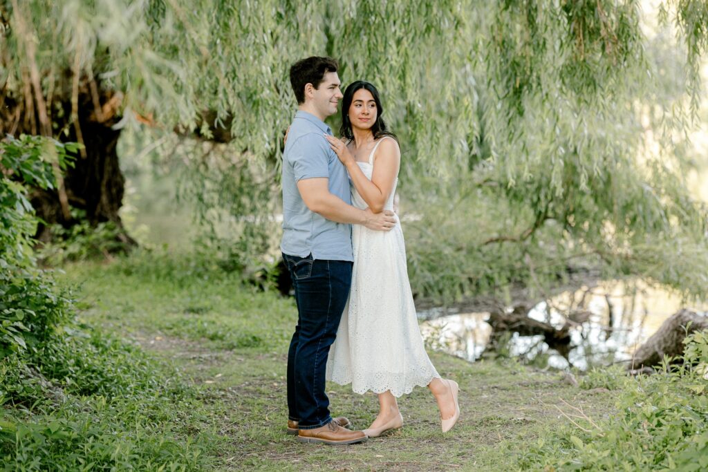 Engaged couple embracing under weeping willow trees at Riverside Park in Grand Rapids