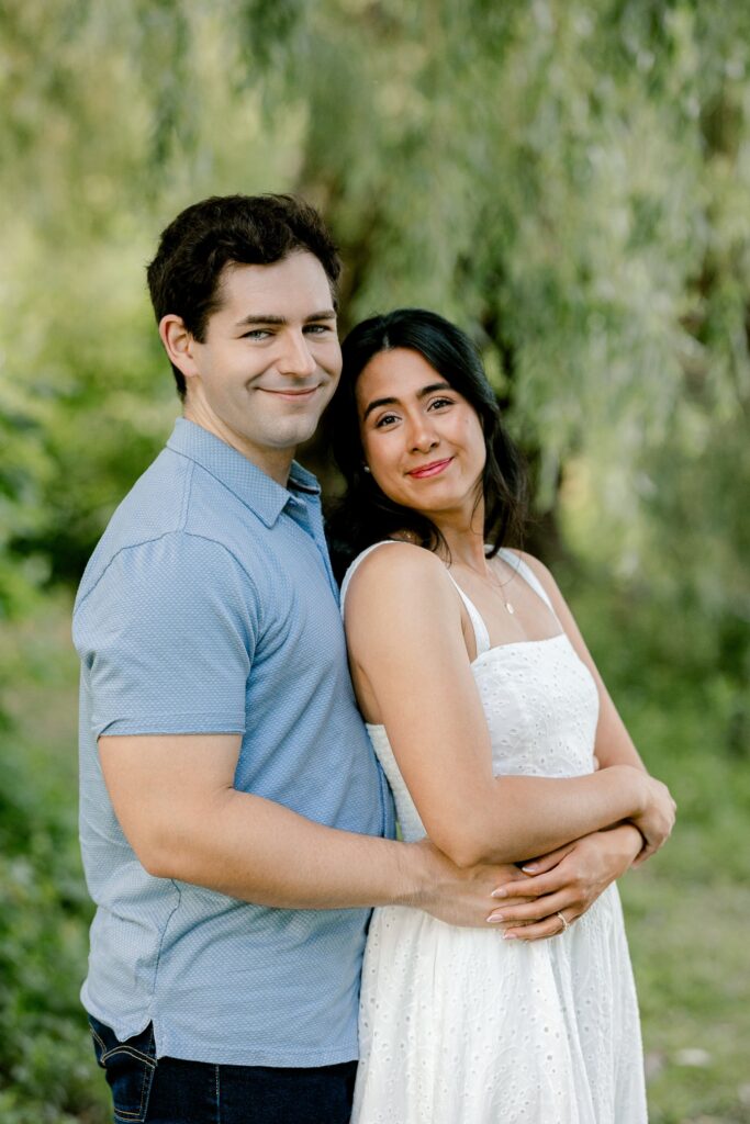 Smiling couple in summer outfits posing in front of willow trees at Riverside Park