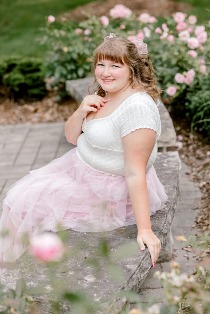 Smiling senior girl sitting among pink roses for her Grand Rapids senior pictures