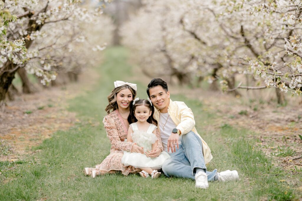 Family sitting close together in the grass between rows of flowering apple trees in Grand Rapids