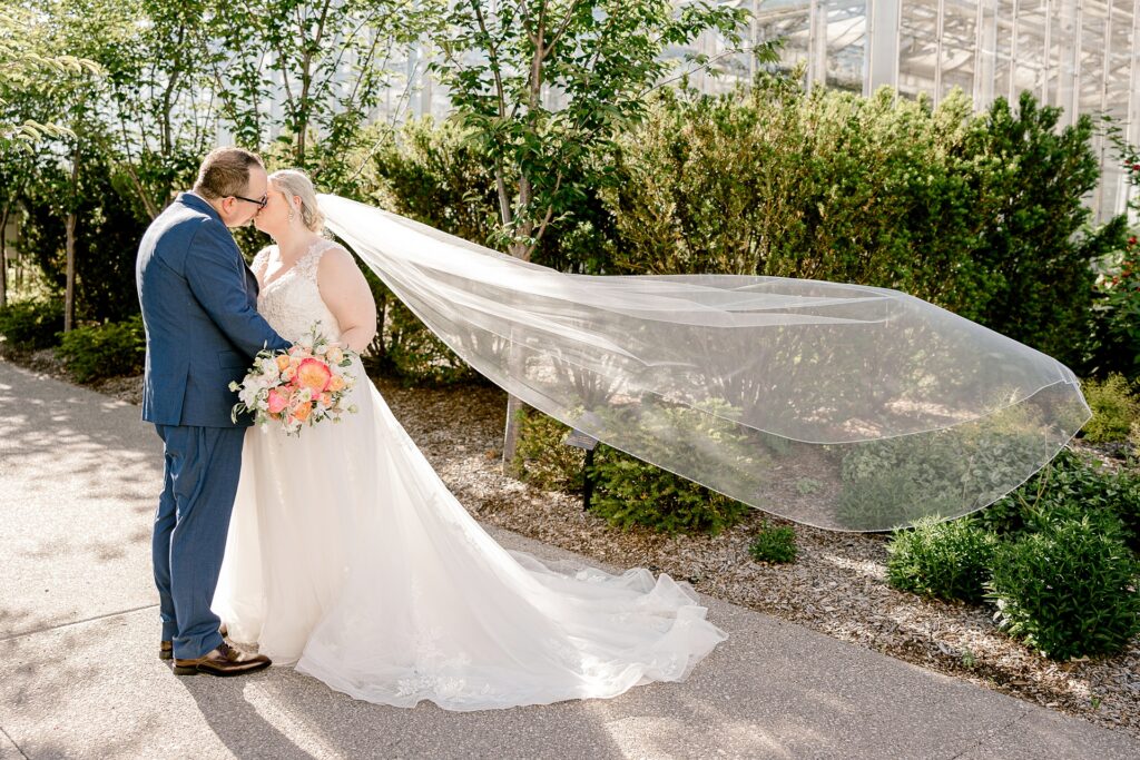 Bride and groom kissing as her veil blows in the breeze at Frederik Meijer Gardens