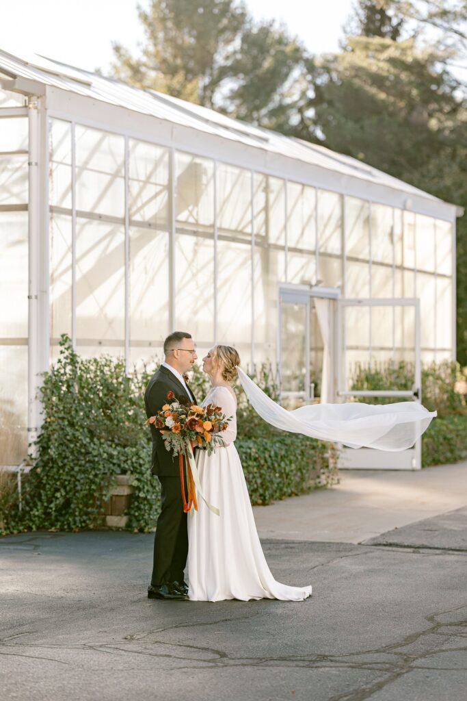 Bride's veil blowing in the wind as the couple stands outside the greenhouse