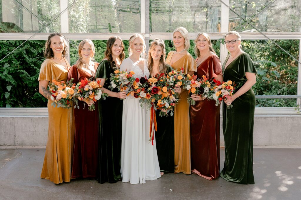 Bridesmaids in rust, mustard and green velvet dresses holding fall bouquets in the greenhouse